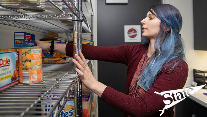 Student retrieving a box of macaroni from a food pantry shelf.