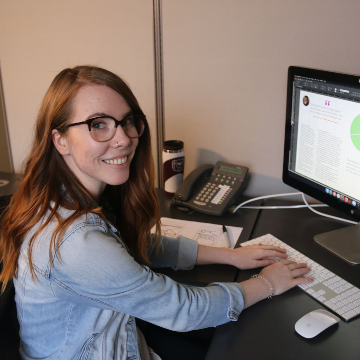 A student with long red hair and gold glasses smiles while working at a computer