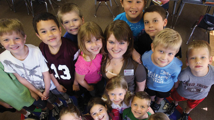A teacher in a classroom surrounded by smiling students.