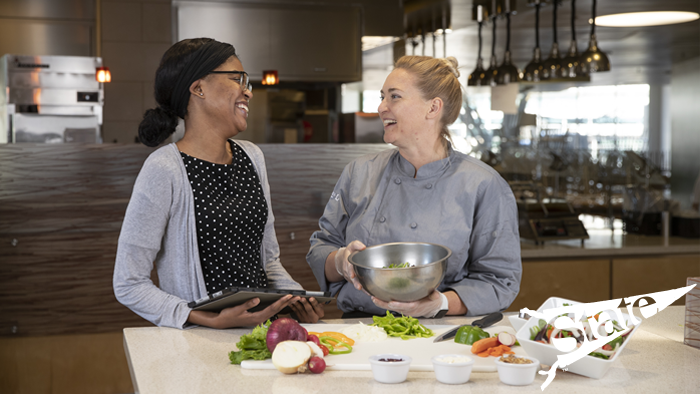 Two women cutting vegetables, looking at each other and smiling